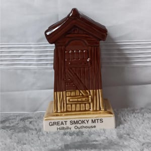 May include: A brown ceramic outhouse figurine with a peaked roof and a wooden plank texture. The base is cream-colored and features the black printed text GREAT SMOKY MTS Hillbilly Outhouse. The structure has a diagonal door brace detail.