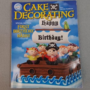 May include: The 2010 Wilton Yearbook Cake Decorating magazine cover features a brown pirate ship cake with blue icing waves, five pirate figurines, and the text: Cake Decorating, Special Section: First Birthday Bash!, Happy Birthday!