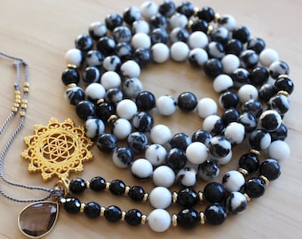 Pode incluir: Um colar mala de pedra marmoreada preto e branco com um pingente dourado de flor da vida e uma gota de cristal de quartzo transparente. O colar é feito com 108 contas e tem uma borla cinza.