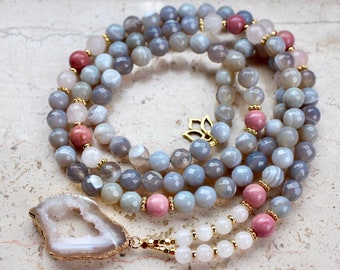 May include: A gray and pink agate gemstone mala necklace with a large, white agate pendant. The necklace features a gold lotus charm and is strung on a gold cord.