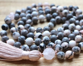Pode incluir: Um colar de contas com contas de pedra cinzentas, brancas e rosa. O colar tem uma borla rosa e uma pedra rosa redonda grande. As contas são enfiadas num cordão, criando um efeito em camadas.