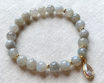 Pode incluir: Um bracelete de pedras cinzas com contas douradas e um pingente de cristal em forma de lágrima.