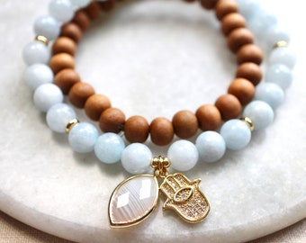 May include: Two bracelets, one with light blue beads and one with brown wooden beads, are stacked on top of each other. A gold charm with a hamsa hand and a white stone hangs from the bracelets.
