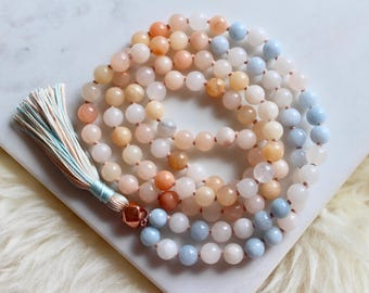 Pode incluir: Um colar mala de pedras preciosas multicoloridas com um tassel azul claro. O colar é feito de contas azul claro, pêssego e brancas.