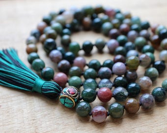 Pode incluir: Um colar mala de pedras preciosas verdes, marrons e rosa com um detalhe de contas turquesa e vermelhas e um tassel azul-esverdeado.