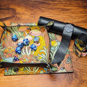 May include: A colorful dice tray with a mushroom and floral design. The tray is made of leather and has a black leather strap. There are blue dice inside the tray.