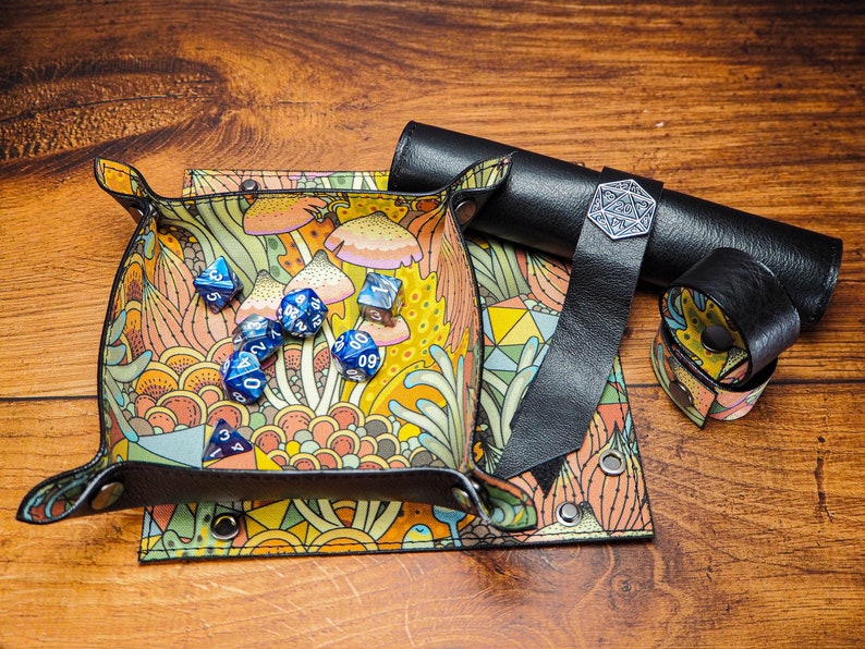 May include: A colorful dice tray with a mushroom and floral design. The tray is made of leather and has a black leather strap. There are blue dice inside the tray.