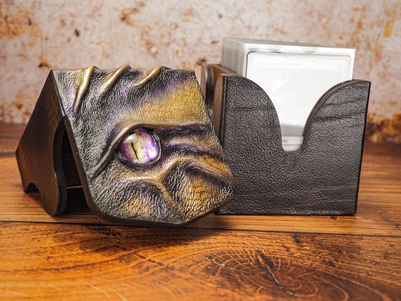 May include: A black leather card box with a dragon design. The box has a purple gem eye and gold accents. The box is open and a stack of cards is visible inside.