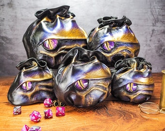May include: Five leather dice bags with a gold and purple iridescent finish. Each bag has a large purple eye design on the front. The bags are sitting on a wooden surface with several purple dice scattered around them.