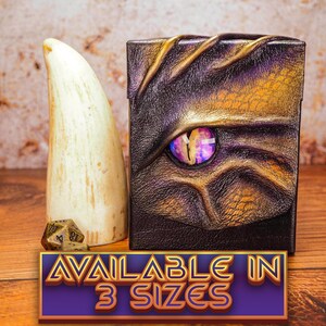 May include: A purple and gold leather dice box with a dragon eye design. The box is closed and sitting on a wooden surface. The text "AVAILABLE IN 3 SIZES" is printed on a purple and gold banner below the box.