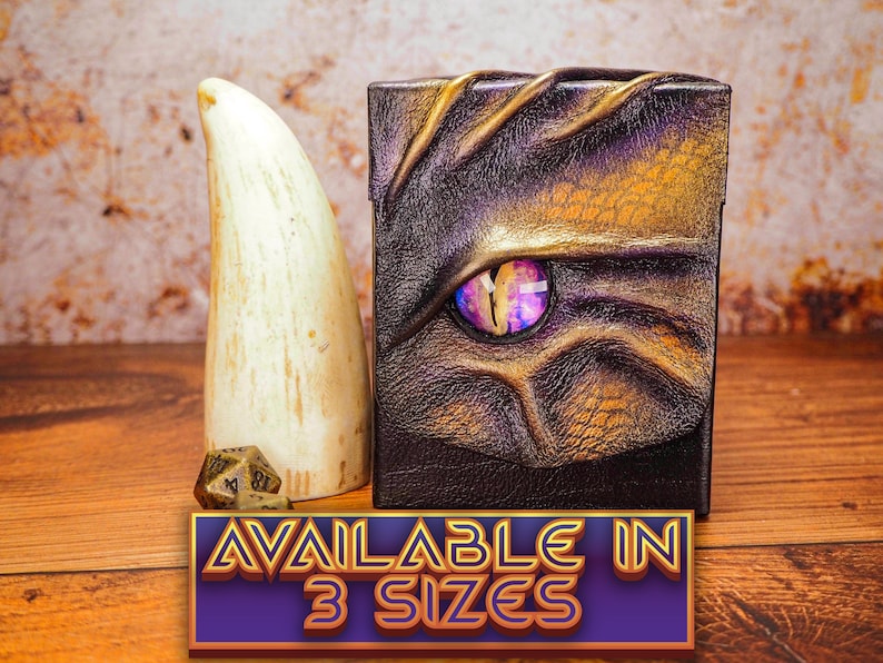 May include: A purple and gold leather dice box with a dragon eye design. The box is closed and sitting on a wooden surface. The text "AVAILABLE IN 3 SIZES" is printed on a purple and gold banner below the box.