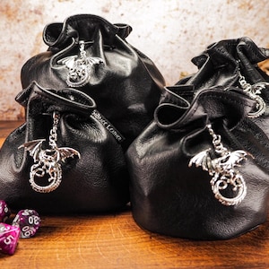 Pode incluir: Quatro sacos de dados de couro preto com pingentes de dragão prateados. Os sacos estão dispostos em forma de pirâmide sobre uma superfície de madeira. Há vários dados roxos espalhados ao redor dos sacos.