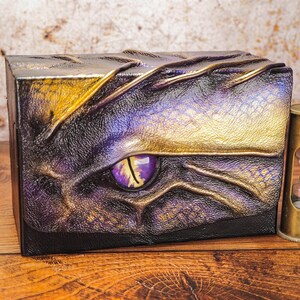 May include: A small, decorative box with a dragon eye design. The box is made of leather and has a gold and purple color scheme. The eye is a vibrant purple with a yellow iris. The box is sitting on a wooden surface.
