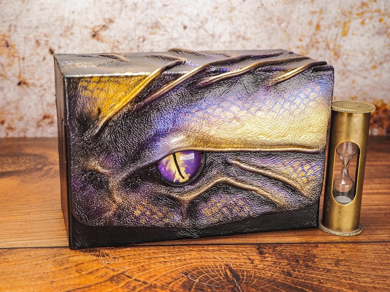May include: A small, decorative box with a dragon eye design. The box is made of leather and has a gold and purple color scheme. The eye is a vibrant purple with a yellow iris. The box is sitting on a wooden surface.