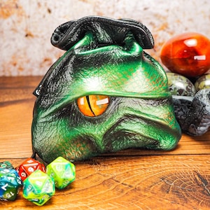 May include: Green leather dice bag with a dragon eye design. The bag is sitting on a wooden table with dice scattered around it.