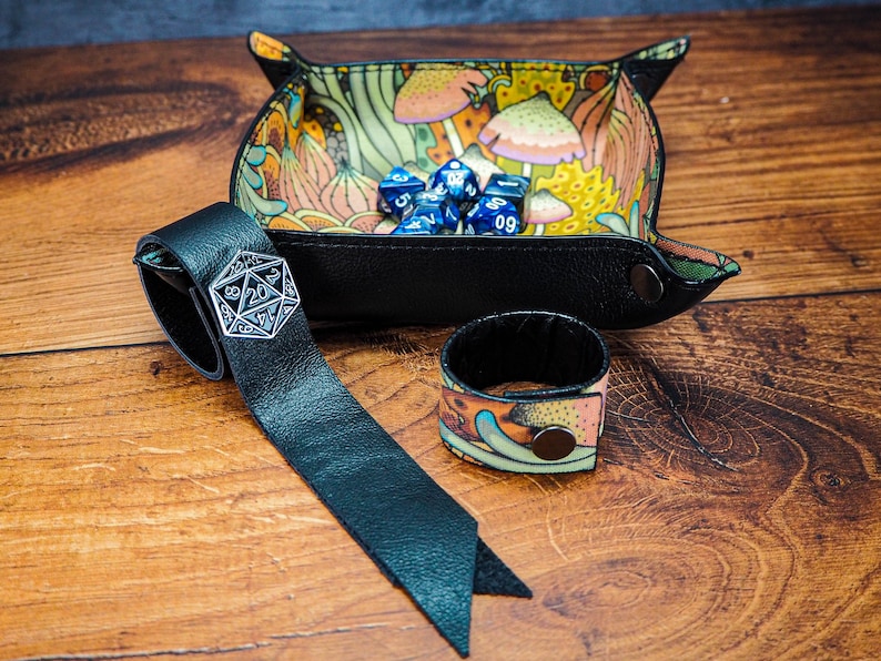 May include: A black leather dice tray with a colorful mushroom design. The tray is filled with blue dice. A black leather dice bag with a silver metal dice charm is also pictured.