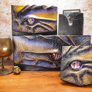 May include: Three leather boxes with dragon eye designs. The boxes are black, purple, and gold with a purple eye in the center of each. The boxes are on a wooden surface with a gold goblet and a dice.