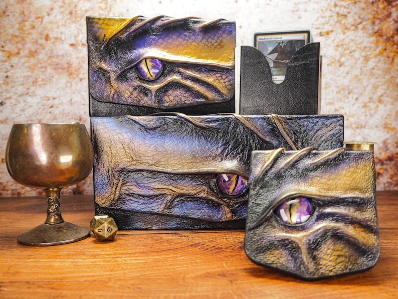 May include: Three leather boxes with dragon eye designs. The boxes are black, purple, and gold with a purple eye in the center of each. The boxes are on a wooden surface with a gold goblet and a dice.