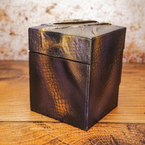 May include: A small, square box with a textured, black and gold finish. The box has a unique, almost scaly, pattern.