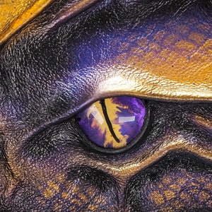 May include: Close-up of a dragon's eye with a purple and gold iris. The eye is set in a textured, leathery surface.