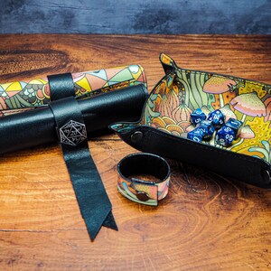 May include: A set of dice rolling accessories for tabletop games. The set includes a black leather dice rolling tray with a colorful floral pattern, a black leather dice rolling tube with a colorful floral pattern, and a black leather dice rolling pouch with a colorful floral pattern. The tray has a metal D20 die on top of it.