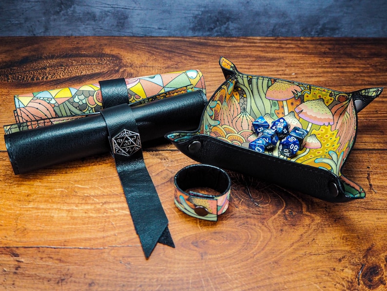 May include: A set of dice rolling accessories for tabletop games. The set includes a black leather dice rolling tray with a colorful floral pattern, a black leather dice rolling tube with a colorful floral pattern, and a black leather dice rolling pouch with a colorful floral pattern. The tray has a metal D20 die on top of it.