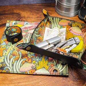 May include: A black leather tray with a colorful mushroom and floral pattern. The tray is filled with various items, including a silver pen, a multi-tool, keys, and coins.