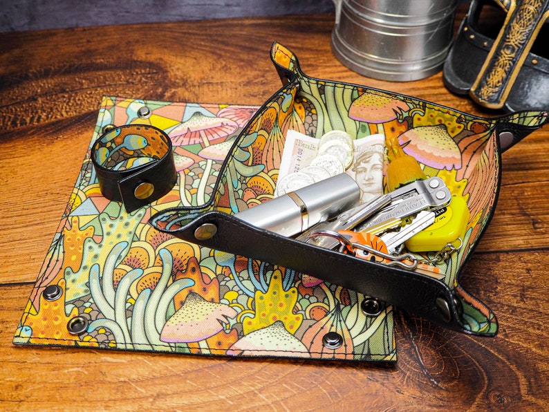 May include: A black leather tray with a colorful mushroom and floral pattern. The tray is filled with various items, including a silver pen, a multi-tool, keys, and coins.