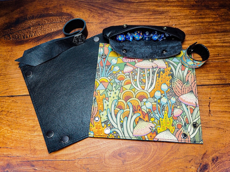 May include: A black leather dice tray with a colorful mushroom and geometric pattern. The tray has a separate compartment for dice, which is lined with black felt. The tray has a black leather wrist strap with a metal buckle.