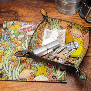 May include: A black leather tray with a colorful mushroom and floral pattern. The tray is filled with various items, including coins, a multi-tool, keys, and a lip balm.