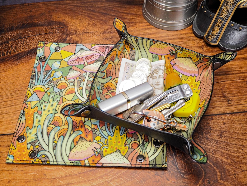 May include: A black leather tray with a colorful mushroom and floral pattern. The tray is filled with various items, including coins, a multi-tool, keys, and a lip balm.