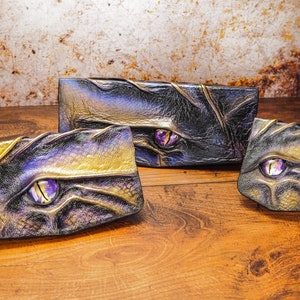 May include: Three leather wallets with a dragon eye design. The wallets are in shades of black, gold, and purple. The eyes are made of glass and have a purple hue.