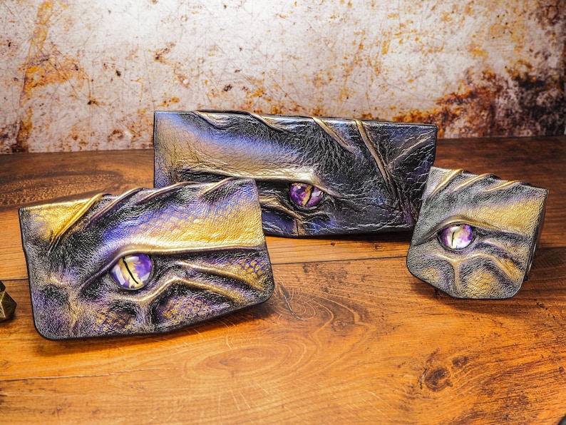 May include: Three leather wallets with a dragon eye design. The wallets are in shades of black, gold, and purple. The eyes are made of glass and have a purple hue.