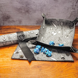 May include: A gray and black dice rolling tray with a variety of fantasy-themed illustrations. The tray is open and has blue dice inside. A matching gray and black dice rolling tube is also included.