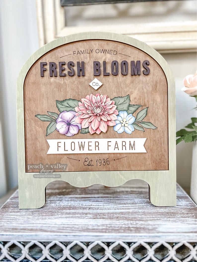 May include: A wooden sign with a brown background and green trim. The sign is shaped like an arch and has the text "FAMILY OWNED. FRESH BLOOMS Co. FLOWER FARM Est. 1936" on it. The sign is decorated with a painted floral arrangement of pink, purple, and blue flowers.