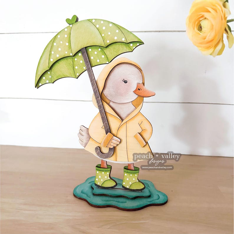 Pode incluir: Estatueta de pato de madeira usando uma capa de chuva amarela e botas de chuva verdes com bolinhas, segurando um guarda-chuva verde com bolinhas. O pato est&aacute; em uma base azul em forma de po&ccedil;a. Com o logotipo peach + valley designs.