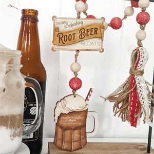 May include: Wooden sign with the text "creamy & refreshing Root Beer Floats" hanging from a beaded garland with red and white beads. A wooden cutout of a root beer float with a straw is hanging below the sign.