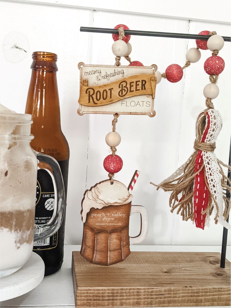 May include: Wooden sign with the text "creamy & refreshing Root Beer Floats" hanging from a beaded garland with red and white beads. A wooden cutout of a root beer float with a straw is hanging below the sign.