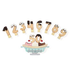 May include: Wooden ice cream cone shaped numbers 1 through 9 and 50. The numbers are decorated with chocolate and sprinkles. The numbers are arranged in a semi-circle with the 50 in the center. The 50 is on top of a banana split with three scoops of ice cream.