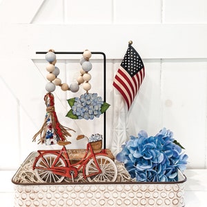 May include: A white metal tray with a red bicycle, blue hydrangeas, a beaded garland, and a small American flag. The tray has a white background with a pattern of small circles.