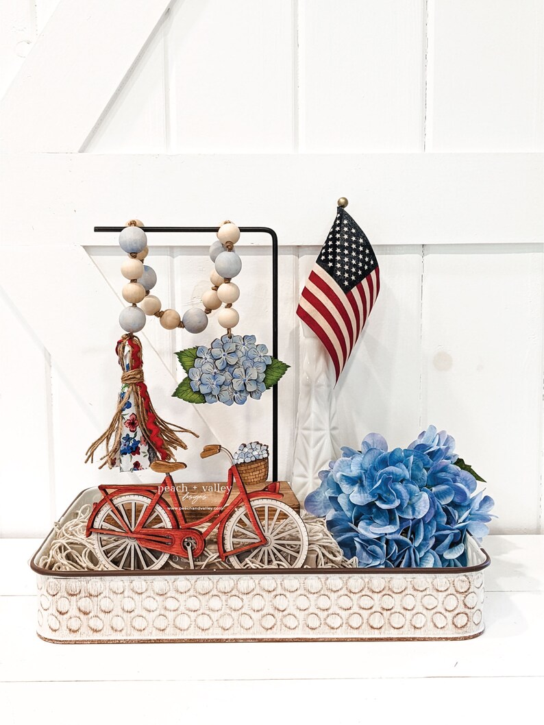 May include: A white metal tray with a red bicycle, blue hydrangeas, a beaded garland, and a small American flag. The tray has a white background with a pattern of small circles.
