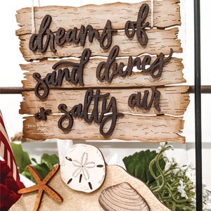 May include: Wooden sign with the text "dreams of sand dunes & salty air". The sign is hanging from a black metal rod. Below the sign is a wooden cutout of a beach scene with a starfish, a seashell, and a sand dollar.