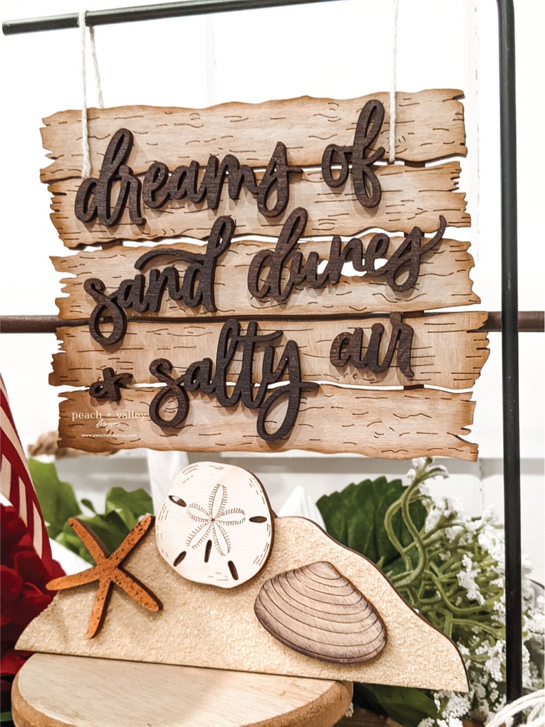 May include: Wooden sign with the text "dreams of sand dunes & salty air". The sign is hanging from a black metal rod. Below the sign is a wooden cutout of a beach scene with a starfish, a seashell, and a sand dollar.