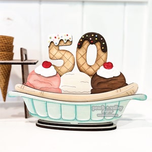 May include: Wooden 50th anniversary decoration shaped like a banana split with three scoops of ice cream, a cherry on top of each scoop, and two waffle cone numbers 5 and 0 with chocolate and sprinkles.