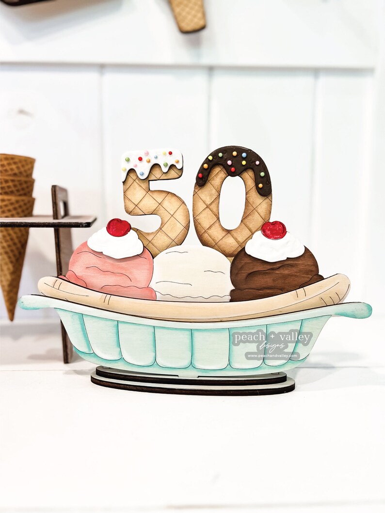 May include: Wooden 50th anniversary decoration shaped like a banana split with three scoops of ice cream, a cherry on top of each scoop, and two waffle cone numbers 5 and 0 with chocolate and sprinkles.