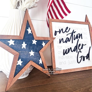 May include: Wooden sign with the text "one nation under God" in black lettering on a white background. The sign is framed in wood with a red, white and blue star in the foreground.