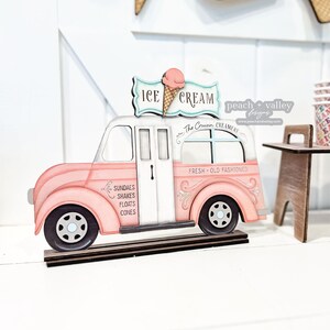 May include: A pink wooden ice cream truck with a white roof and a sign that says "ICE CREAM" with a pink ice cream cone on top. The truck has a window that says "The Crusin CREAMERY" and another window that says "FRESH OLD FASHIONED". The truck has a side panel that says "SUNDAES SHAKES FLOATS CONES".