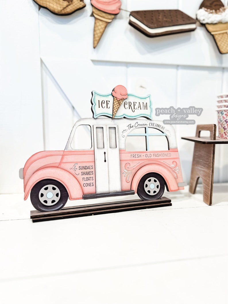 May include: A pink wooden ice cream truck with a white roof and a sign that says "ICE CREAM" with a pink ice cream cone on top. The truck has a window that says "The Crusin CREAMERY" and another window that says "FRESH OLD FASHIONED". The truck has a side panel that says "SUNDAES SHAKES FLOATS CONES".