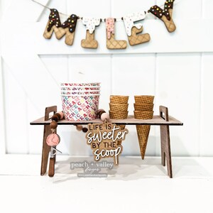 May include: A wooden ice cream stand with a sign that reads "Life is sweeter by the scoop". The stand is decorated with a string of ice cream cone letters that spell "MILEY" and a scoop of ice cream. There are also several ice cream cones and cups on the stand.