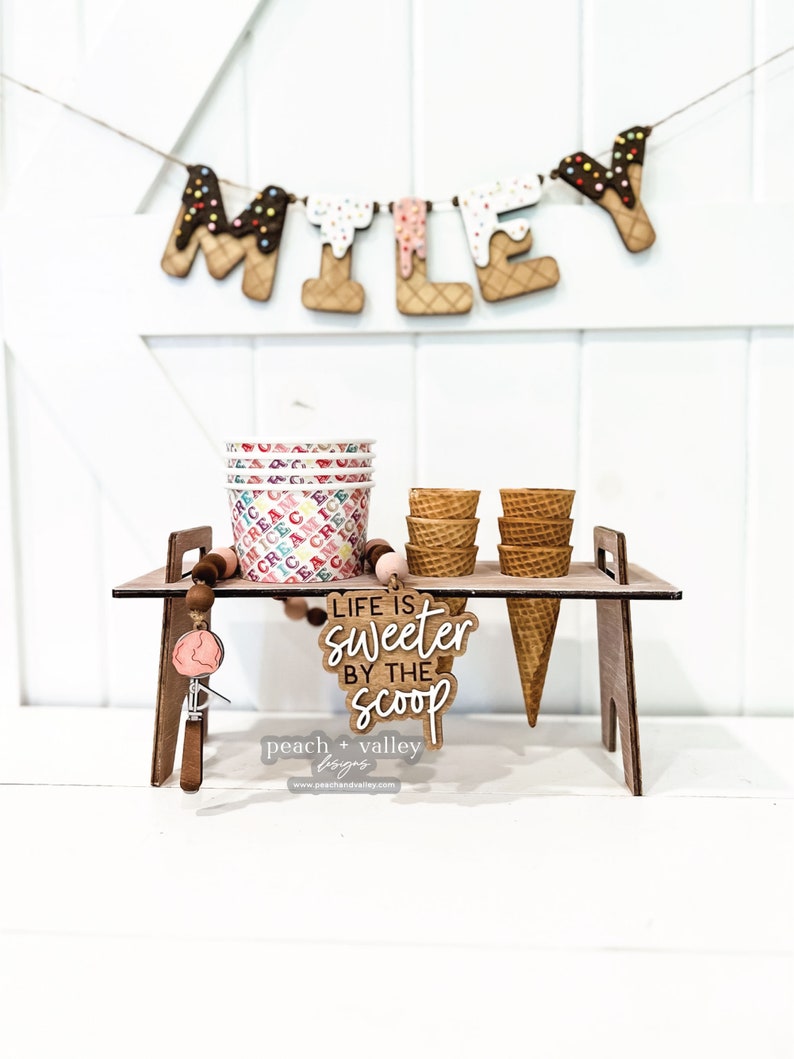 May include: A wooden ice cream stand with a sign that reads "Life is sweeter by the scoop". The stand is decorated with a string of ice cream cone letters that spell "MILEY" and a scoop of ice cream. There are also several ice cream cones and cups on the stand.